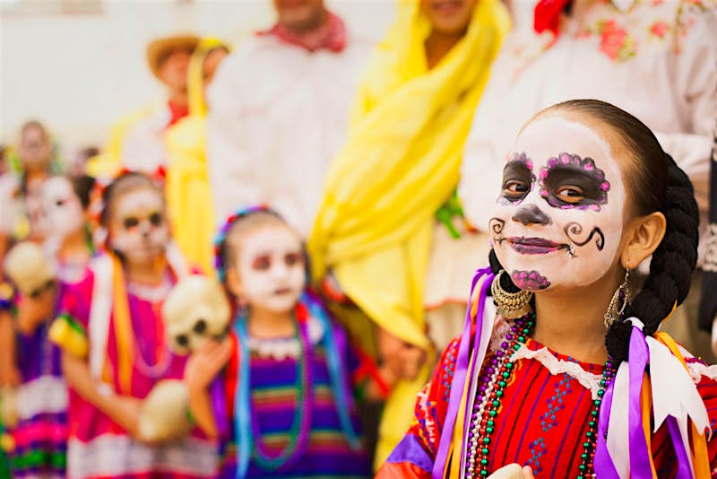 Features - Hero-image-9c87e5081131 A young girl smiles at the camera with her face painted like a calavera and dressed in colorful traditional Mexican clothing