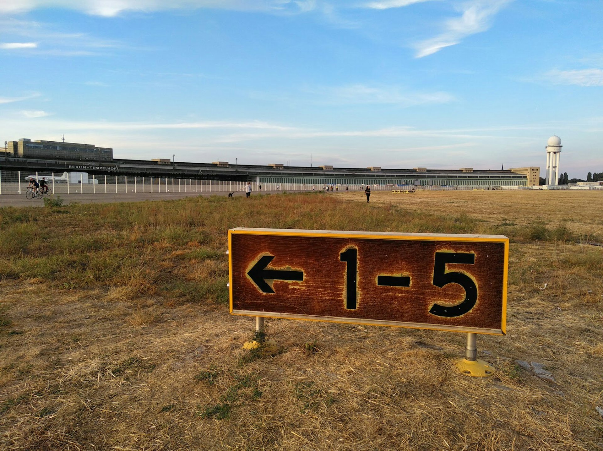 An aged sign at Berlin's abandoned airport, Tempelhofer Feld. The background shows an expanse of dry grass, the old terminal building and people out cycling, jogging, walking dogs and having picnics