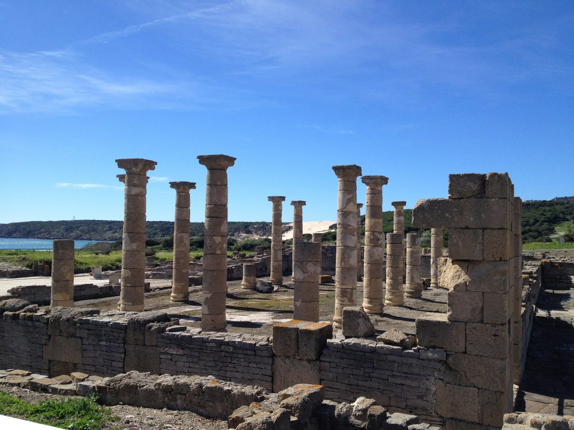 Roman ruins of Baelo Claudia in Bolonia under a clear blue sky. The ruins consist of several intact stone pillars and some half pillars as well as the buildings foundation made out of smaller stones and one corner made from larger, brick-like stones. 