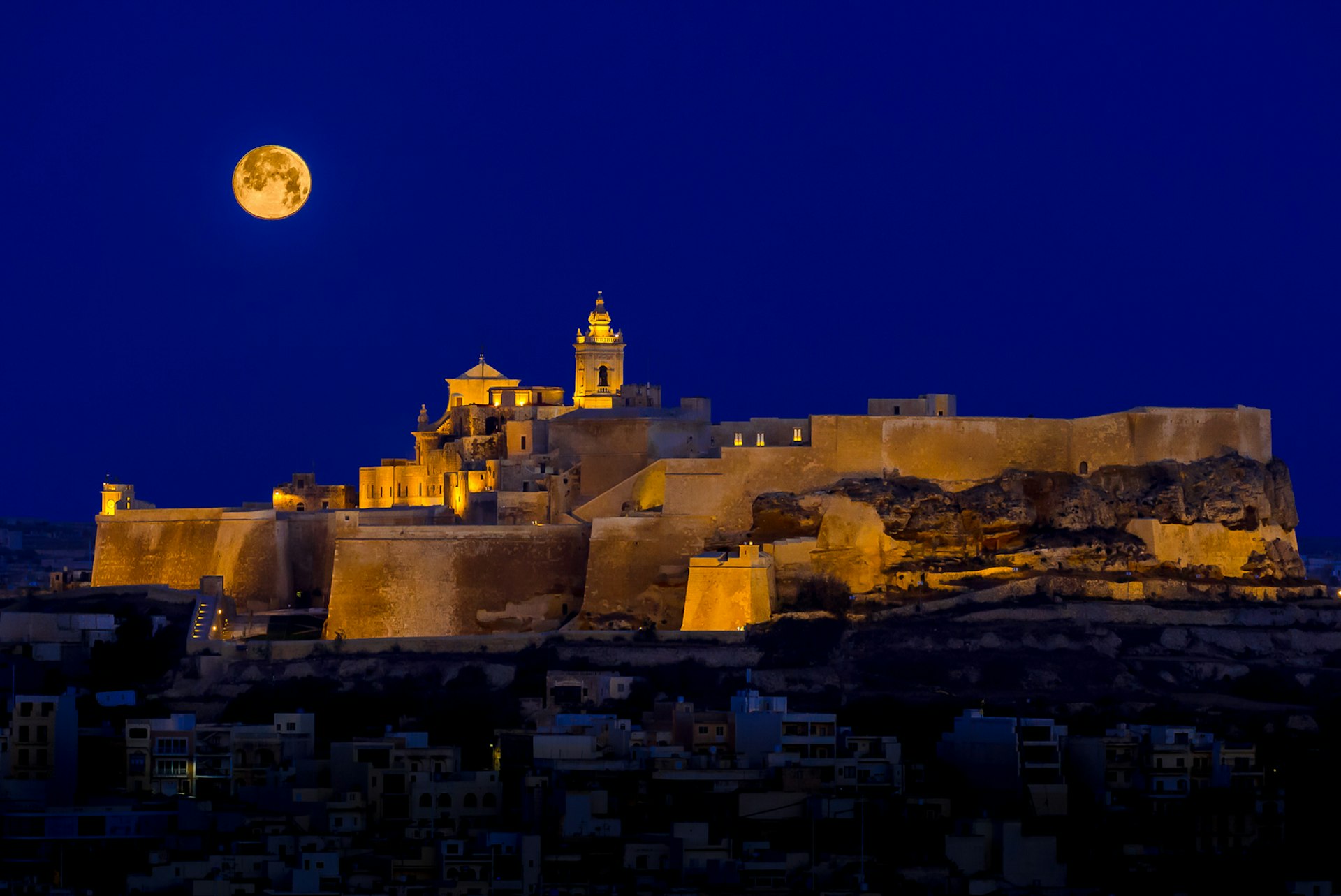 With a full moon rising into a dark sky, the Ċittadella glows gold in floodlights © rossmagri / Getty Images
