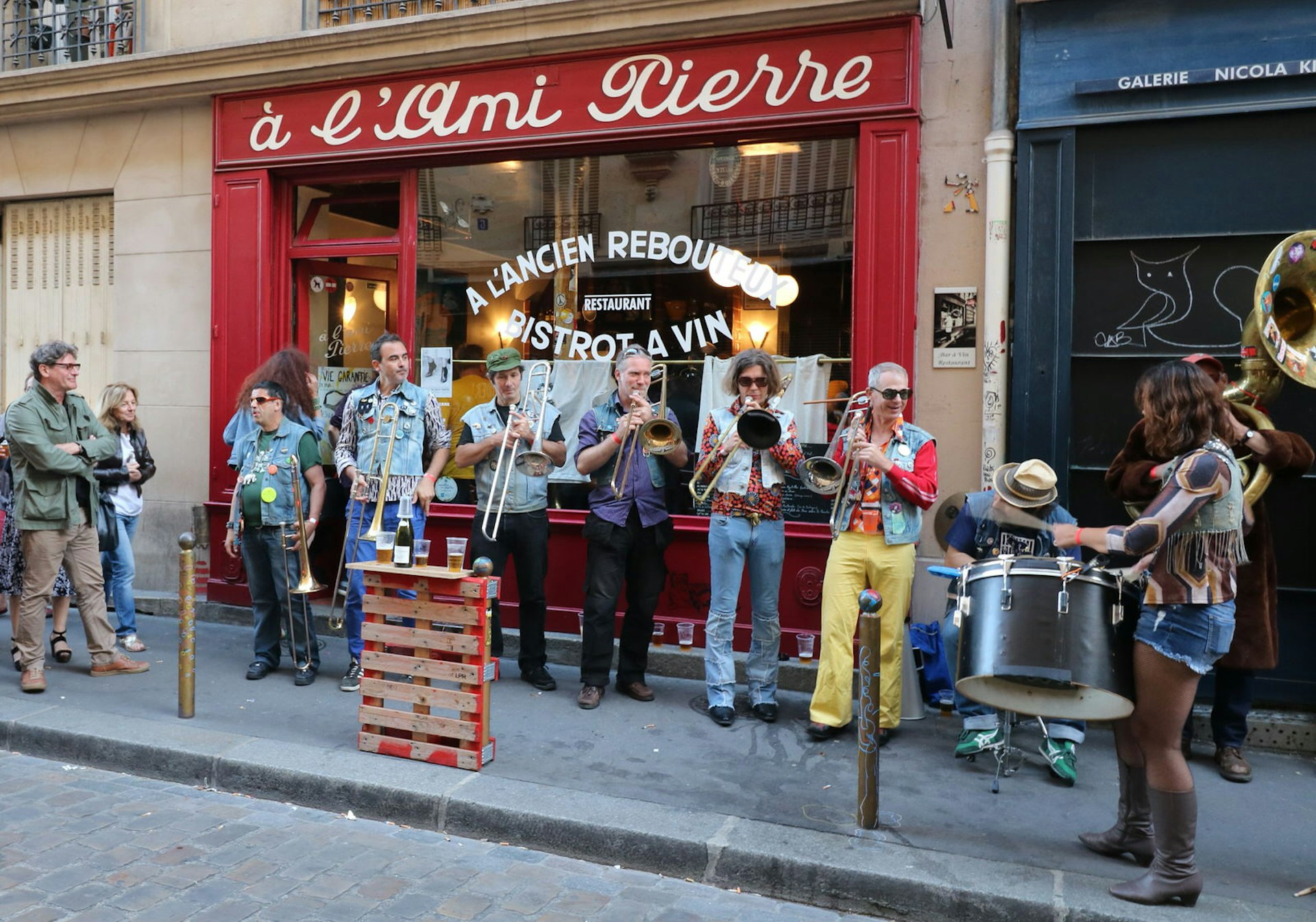 A jazz band, brightly attired in loud shirts and denim, play on the pavement in paris. An upturned shipping pallet is used as a makeshift table, holding glasses of beer and a bottle of wine © Catherine Le Nevez / Lonely Planet