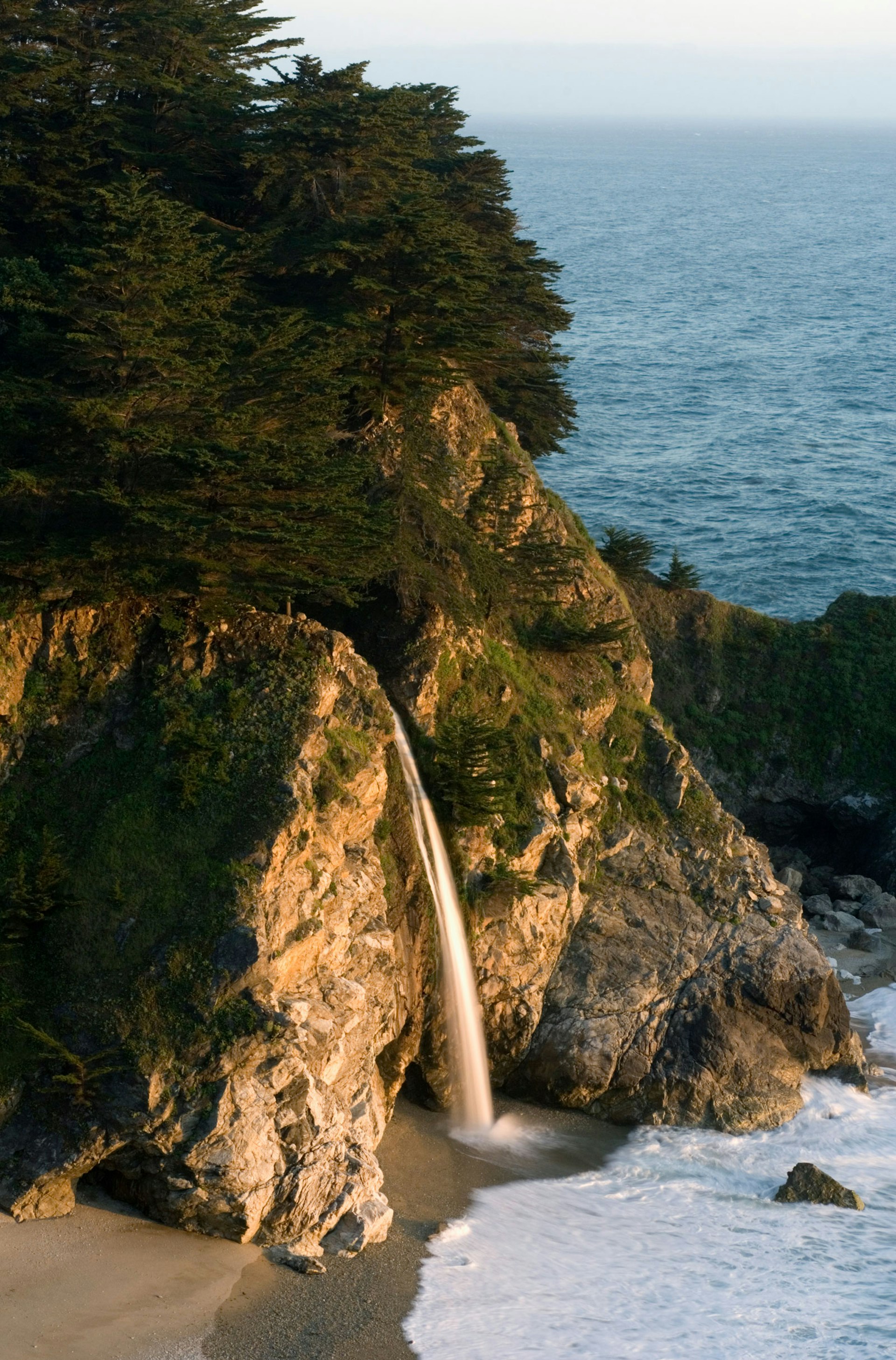 Trees grow on cliffs where a waterfall drops onto the beach near the Pacific Ocean