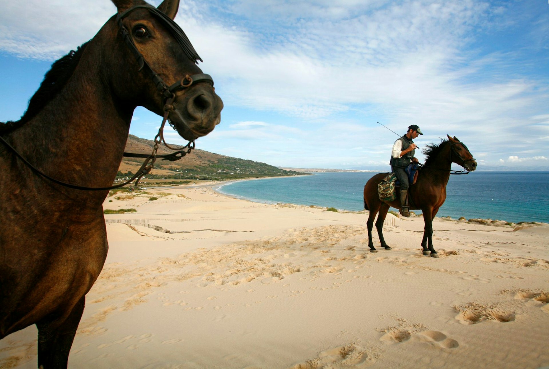 Two people riding brown horses on the otherwise deserted dune of Ensenada de Valdvaqueros with a view over Playa de los Lances beach.