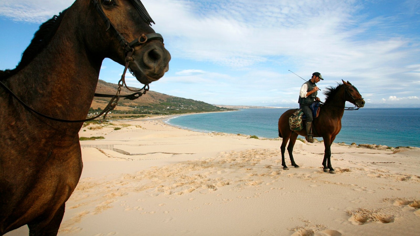 People riding horses on the dune of Ensenada de Valdvaqueros with a view over Playa de los Lances beach.