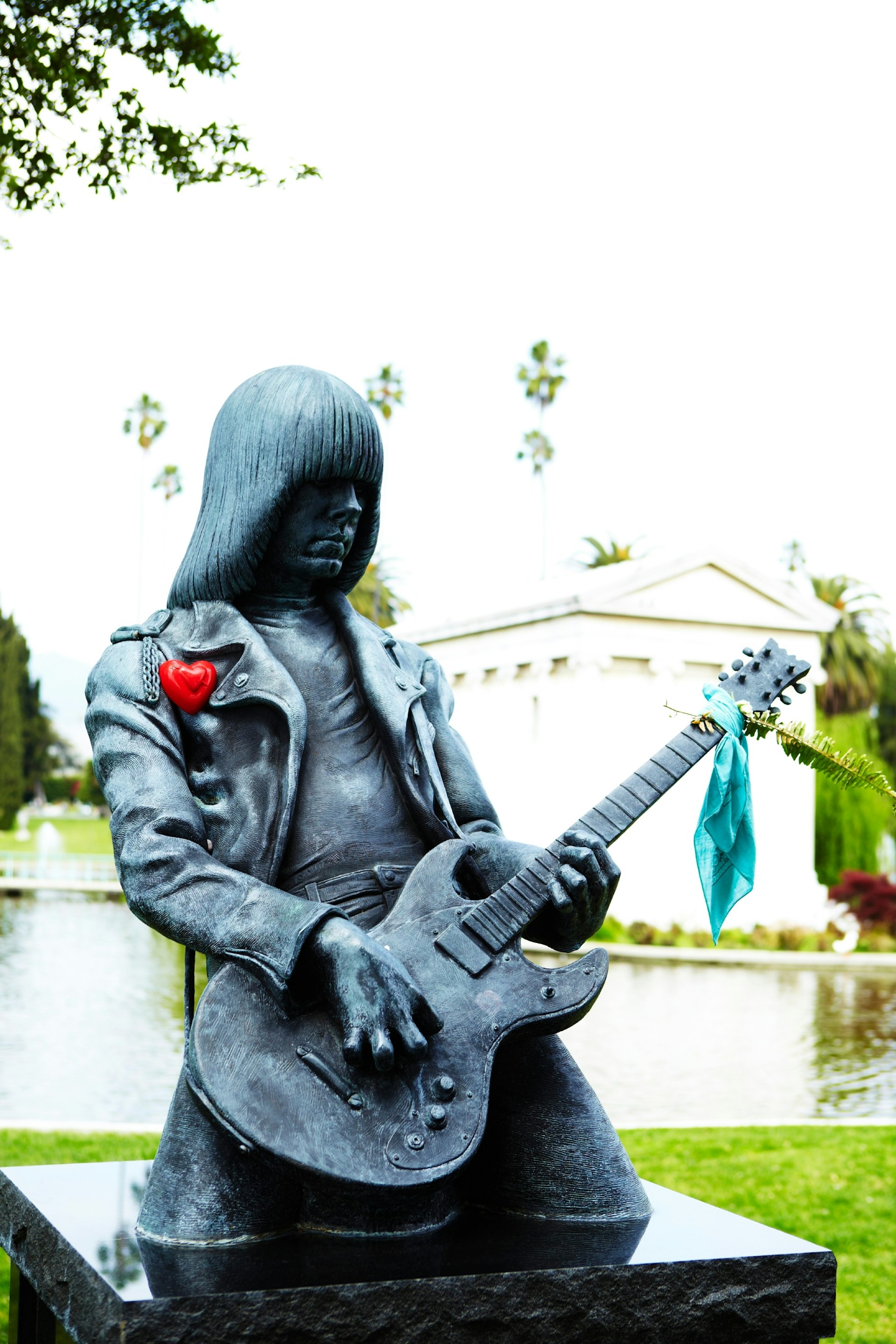 A statue of a long-haired rocker playing the guitar. The guitar has a green scarf tied to it, which holds a piece of fern at the end of the fret board