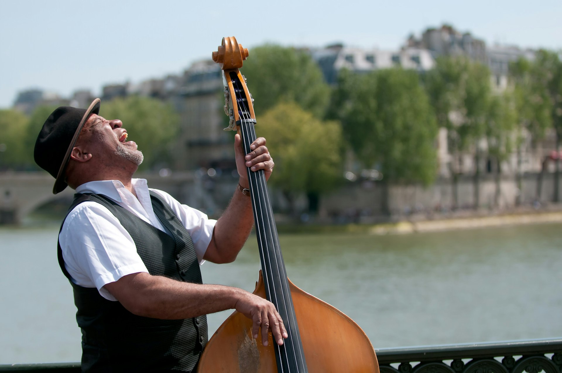 Wearing a trilby-style hat, a street artist playing a double bass has his head tilted back as he belts out a song - eyes closed. In the background is the River Seine. © Christian Rummel / Getty Images