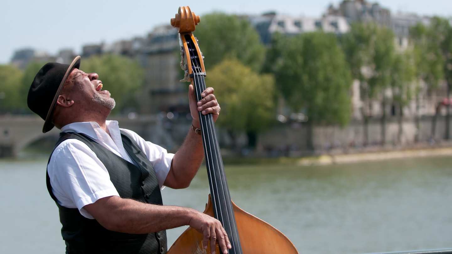 Wearing a trilby-style hat, a street artist playing a double bass has his head tilted back as he belts out a song - eyes closed. In the background is the River Seine. © Christian Rummel / Getty Images