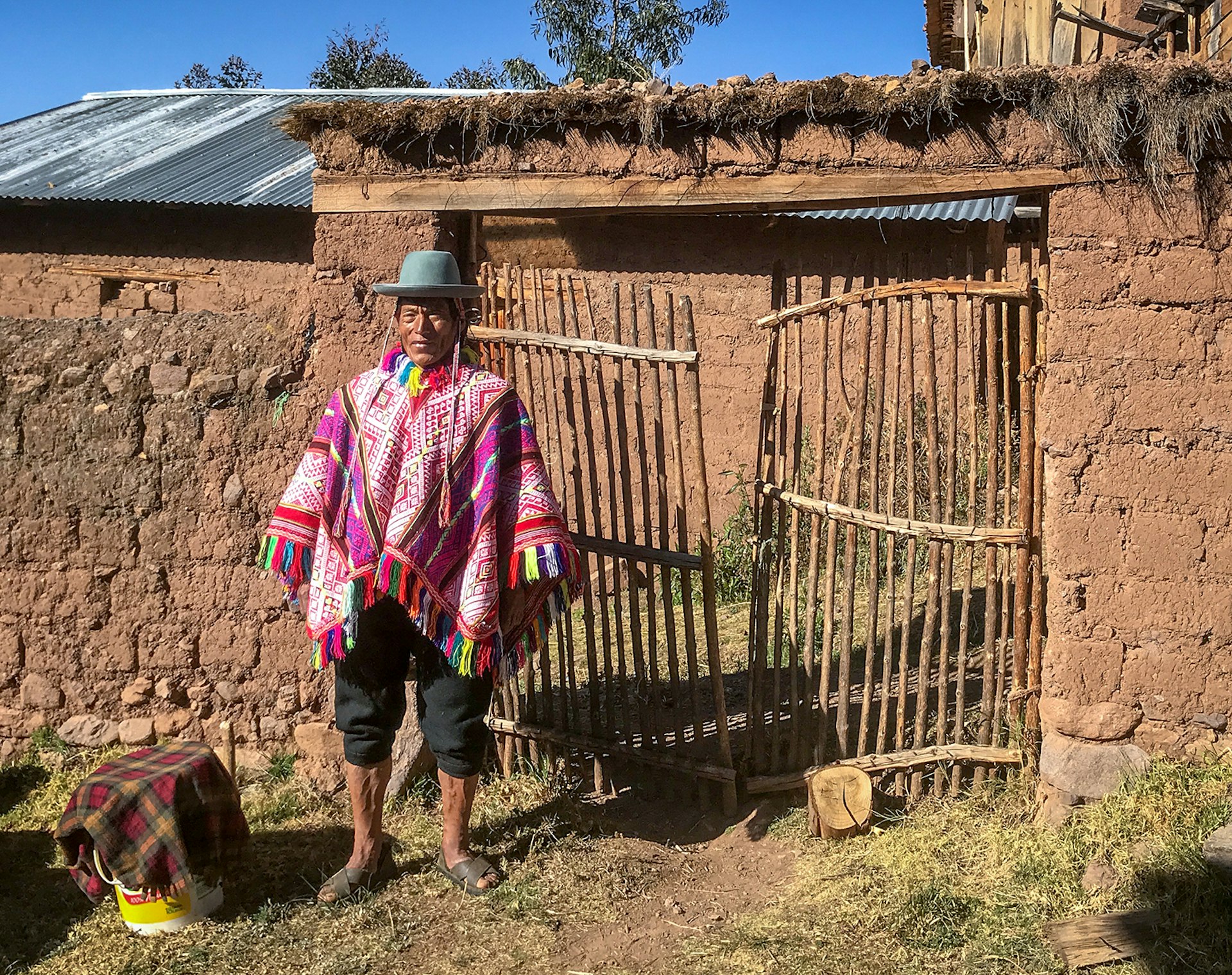 A man in a colorful traditional Andean poncho and hat poses in front of a wall and wooden gate