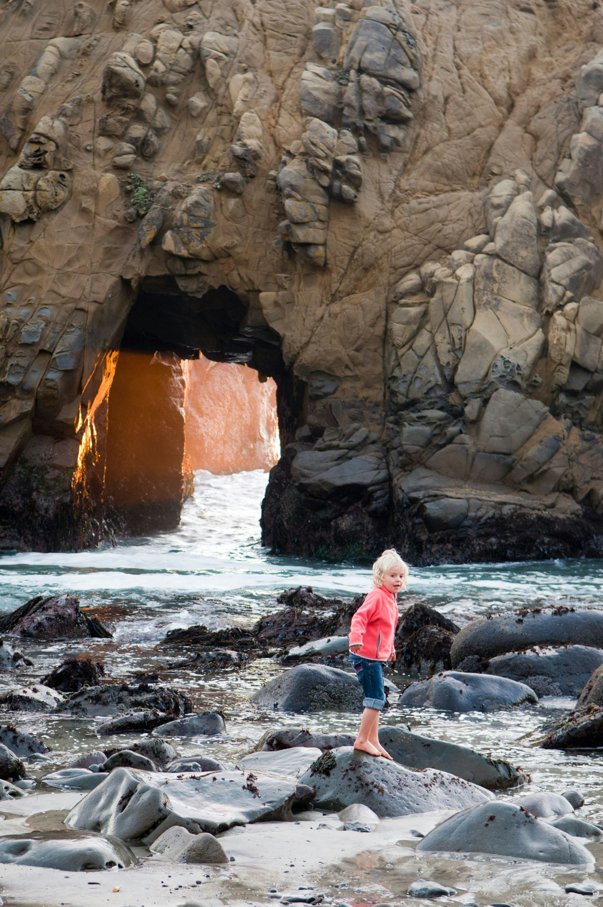 A little girl in a pink coat plays on a rocky coast line with a stone cave behind her