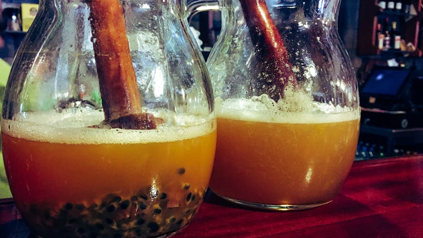 Two carafes with large wooden muddlers inside them on a bartop containing a brown cocktail called poncha, a Madeiran speciality. Passion fruit pips are visible at the bottom of the jugs.