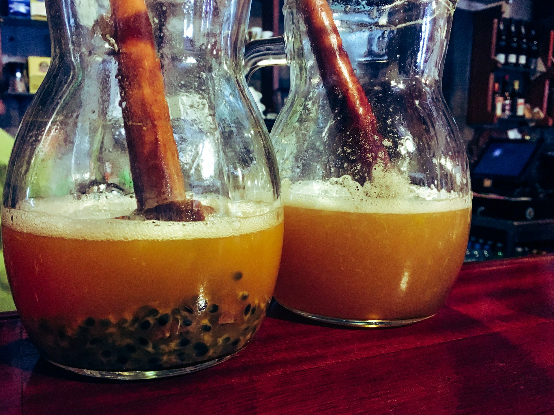 Two carafes with large wooden muddlers inside them on a bartop containing a brown cocktail called poncha, a Madeiran speciality. Passion fruit pips are visible at the bottom of the jugs.