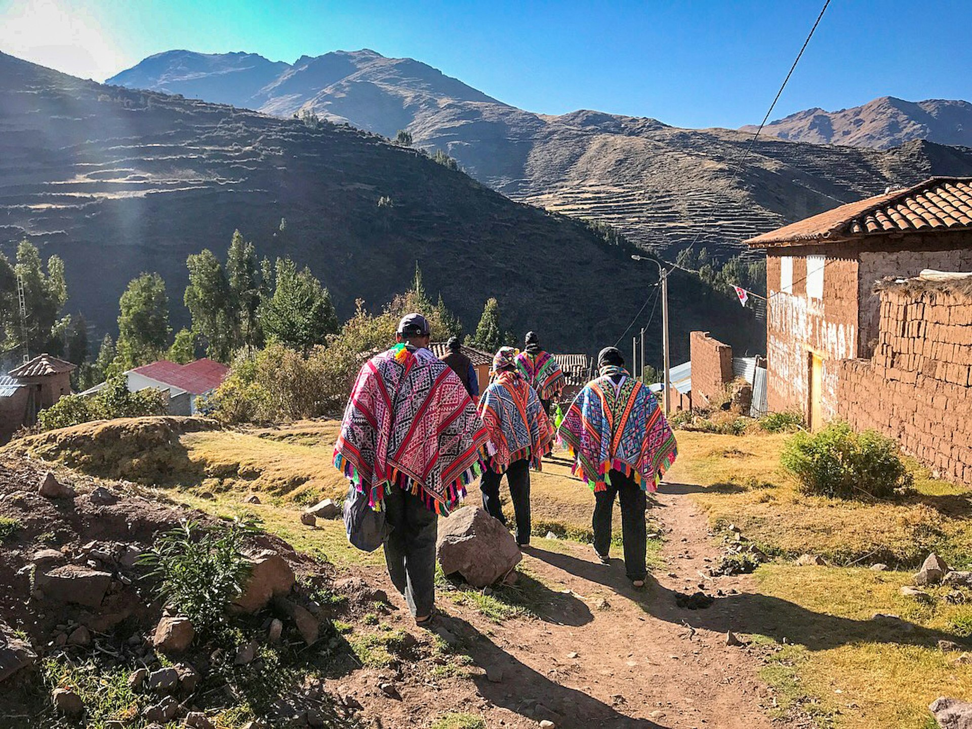 Five men in colorful Andean ponchos walk down a dirt path with a view of mountains in the background