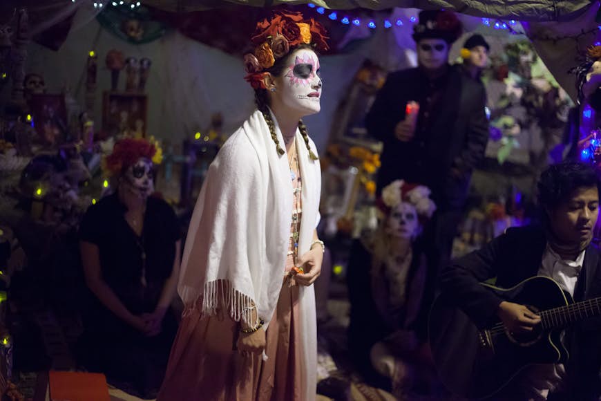 A woman draped in a white shawl with her face painted in the Calavera style sings in front of others dressed for Dia de los Muertos and their ofrendas