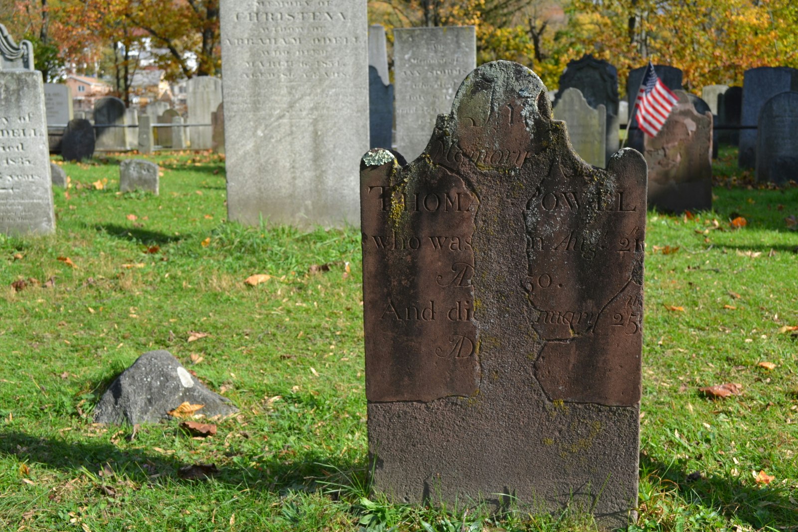 A weathered headstone with faded inscriptions in an old cemetery, with the US flag in the background