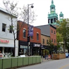 Two green spires rise in the background as several store- and bar-fronts open from colorful buidlings onto a shared patio.