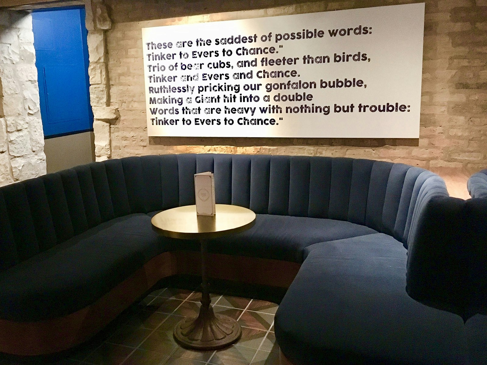 A cornflower blue velvet banquette sits in front of a white brick wall with a large-format reprint of the 1910 poem "Tinker to Evers" on it at the cocktail bar Tinker to Evera