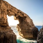 Sunlight bursts through a huge, angular rock arch on the coast of Goza at Wied il-Mielah. Wave surge between the two rock arms © Ramon Portelli / Getty Images