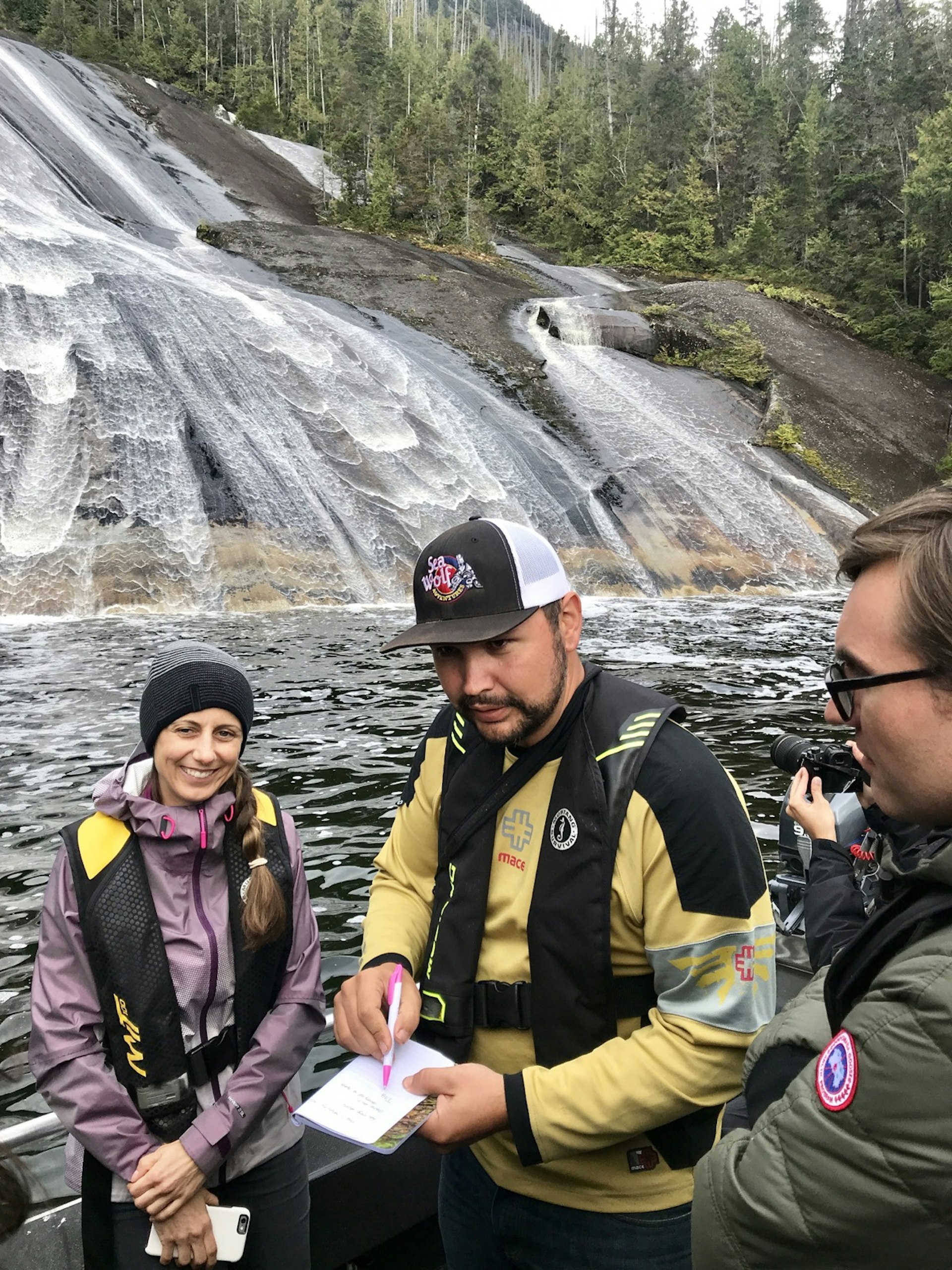 A man is surrounded by travelers on the back deck of a boat as he goes over some interesting points in front of a large waterfall in British Columbia.