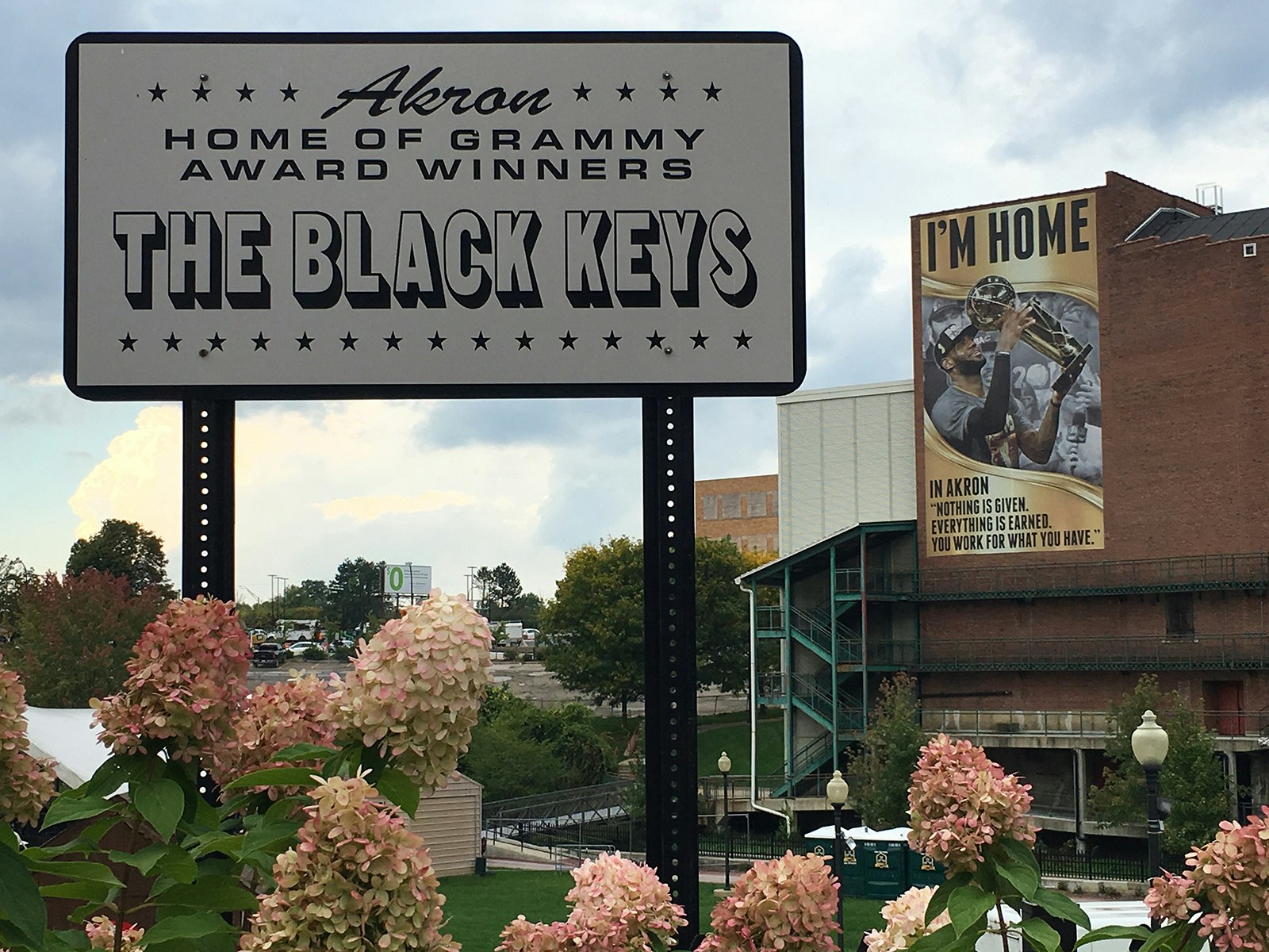 Sign reading "welcome to Akron Ohio, home of the Black Keys' surrounded by hydrangeas with a stadium in the background 