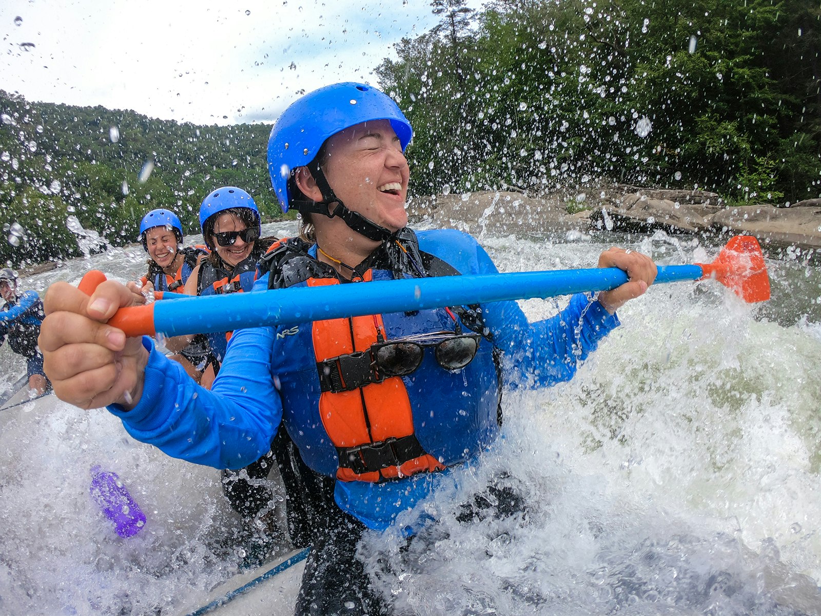 Three women wearing blue and orange life jackets and blue helmets paddle into whitewater on an early fall day on the Gauley River in West Virginia