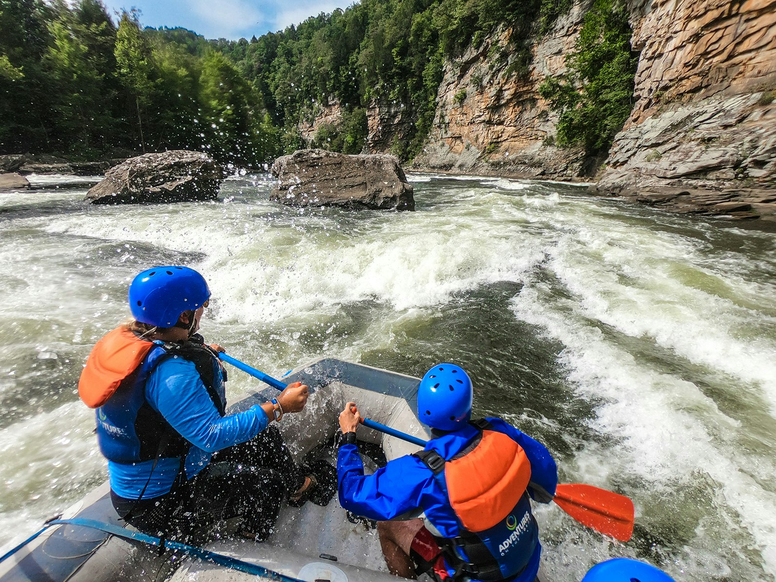 A man and a woman hold paddles at the front of a whitewater raft as they enter a rapid that faces red sandstone cliffs in West Virginia 