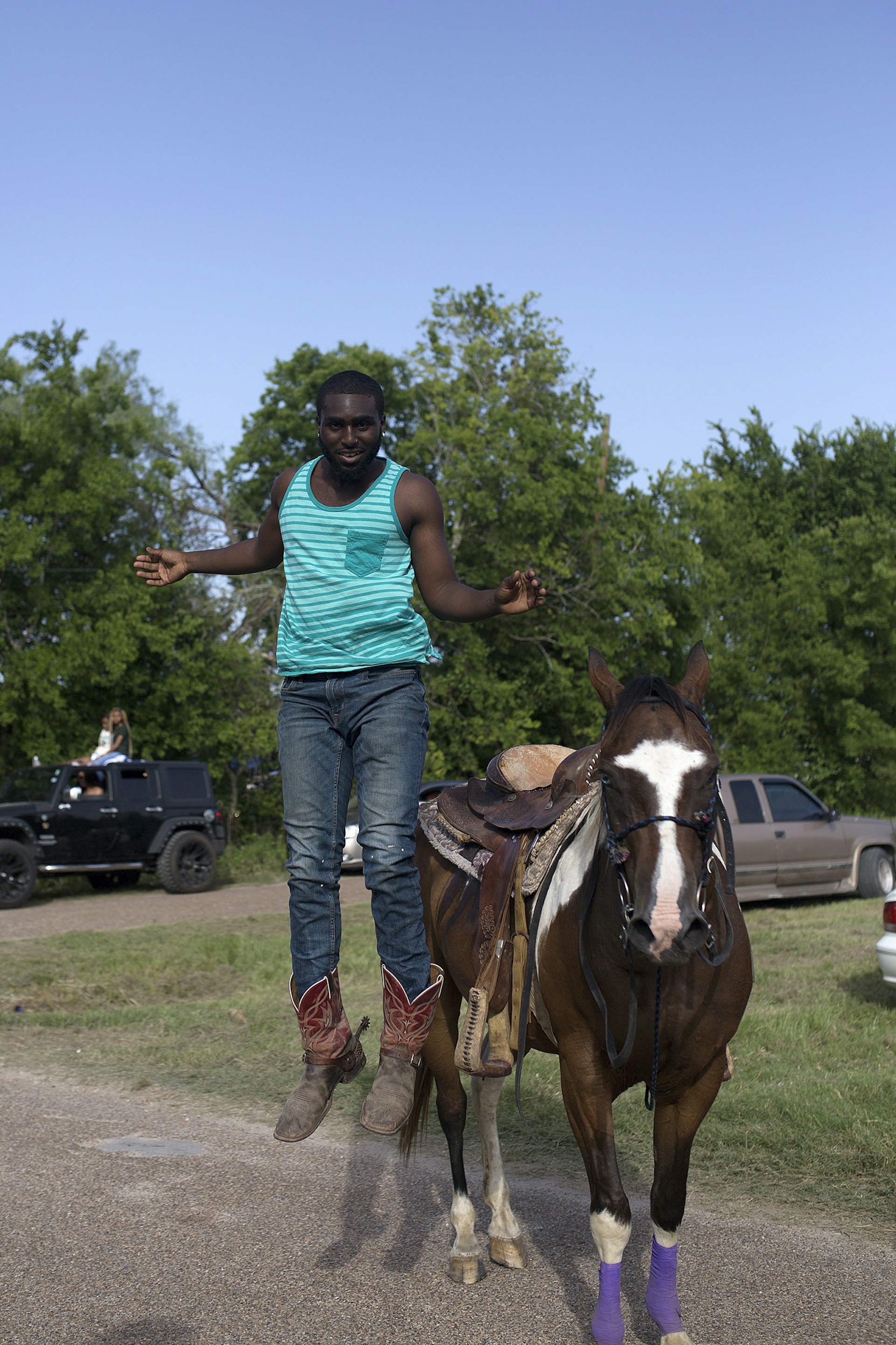 man in a blue tank top, cowboy boots and jeans jumps from the back of a saddled horse with a white blaze on its forehead on a sunny day in the Southern USA