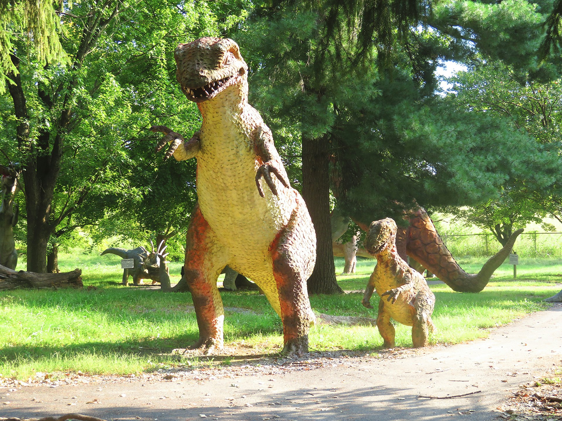 A large fiberglass T Rex is followed by a baby T Rex along a dirt path framed with old trees at a park in the Shenandoah Valley during the summer