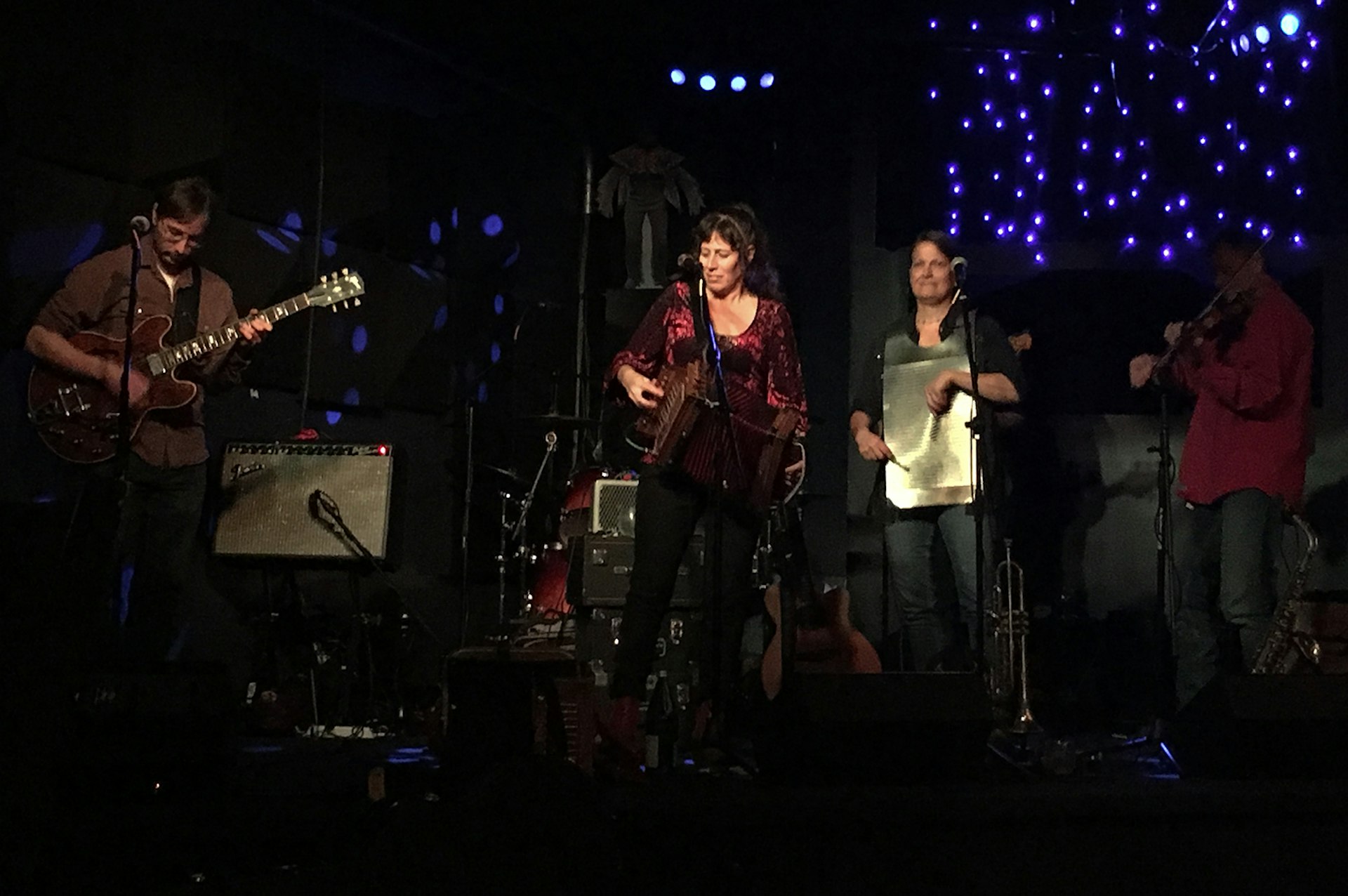 A man with a guitar, a woman singing, a woman with a washboard and a man on a flute perform music on stage in a dark club