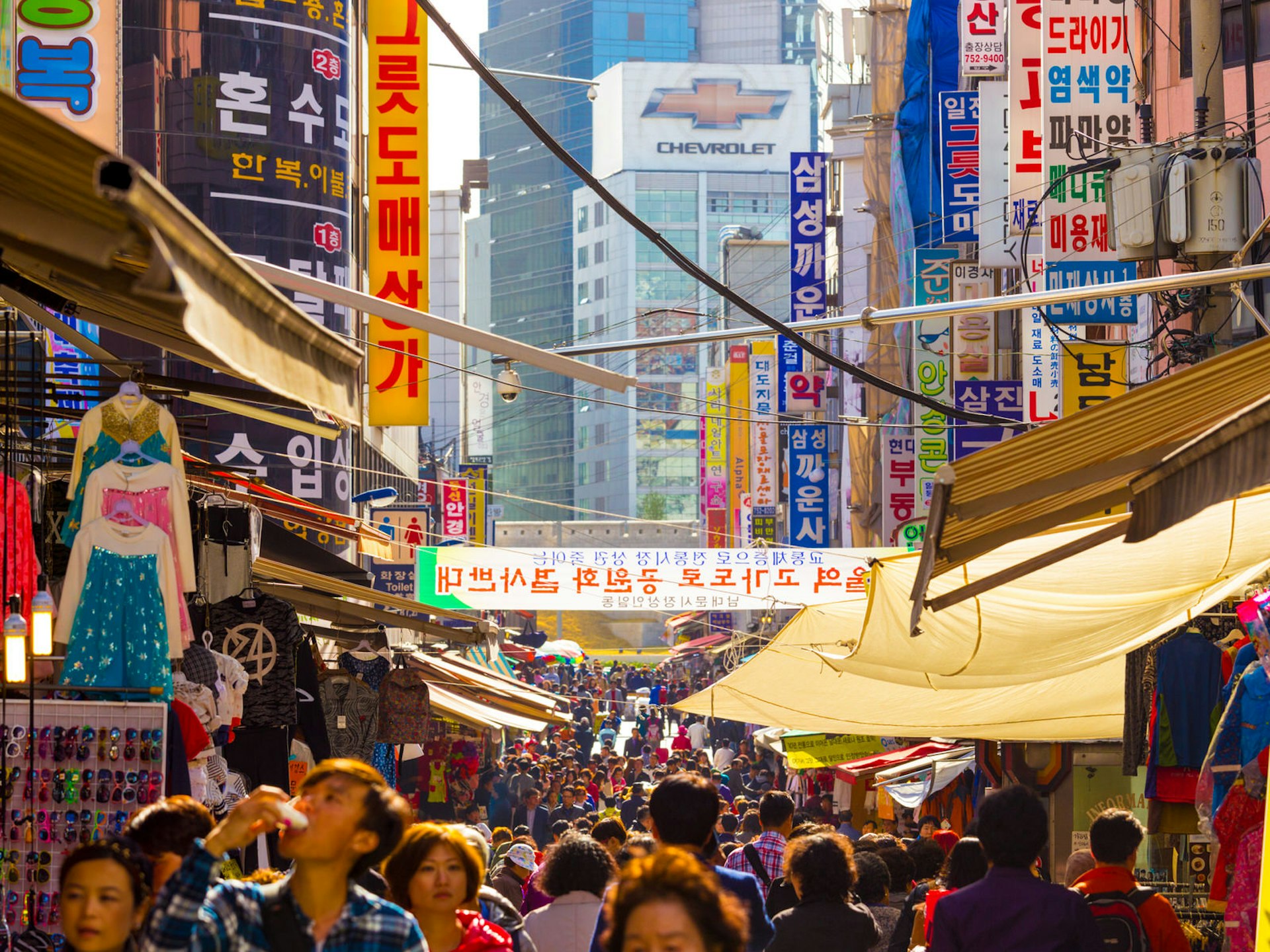 Shoppers fill the main avenue of Namdaemun Market, with stores on both sides and awnings overhanging.