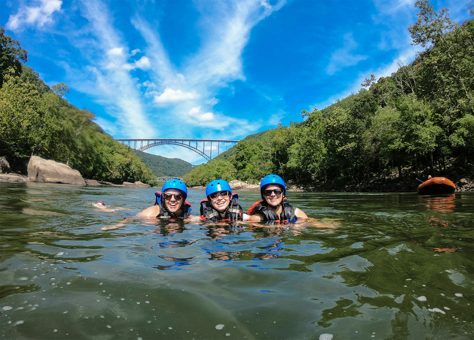 Three women float in a river under a blue sky in a tree-lined gorge. A single-arch bridge spans the river in the background