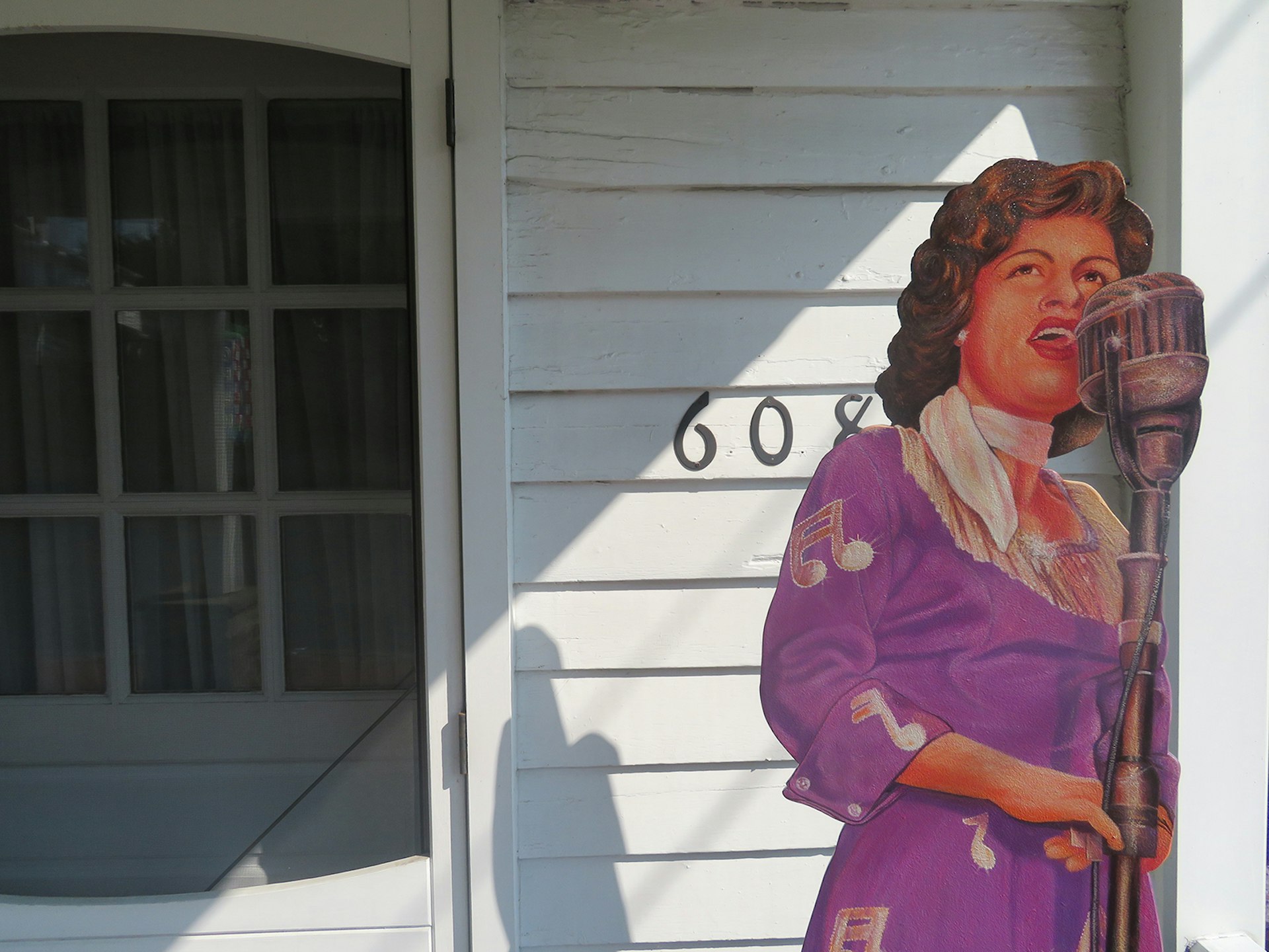 close up of screen door on a white house with the street number '608' and a cardboard cutout of Patsy Cline in a purple dress on the porch