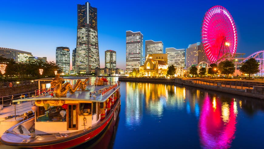 Cityscape of Yokohama city at dusk; in the foreground is the city's port, with an ornate red and gold boat with a dragon on its roof docked in the harbour; in the background are lit-up modern skyscrapers and a huge ferris wheel illuminated with pink lights