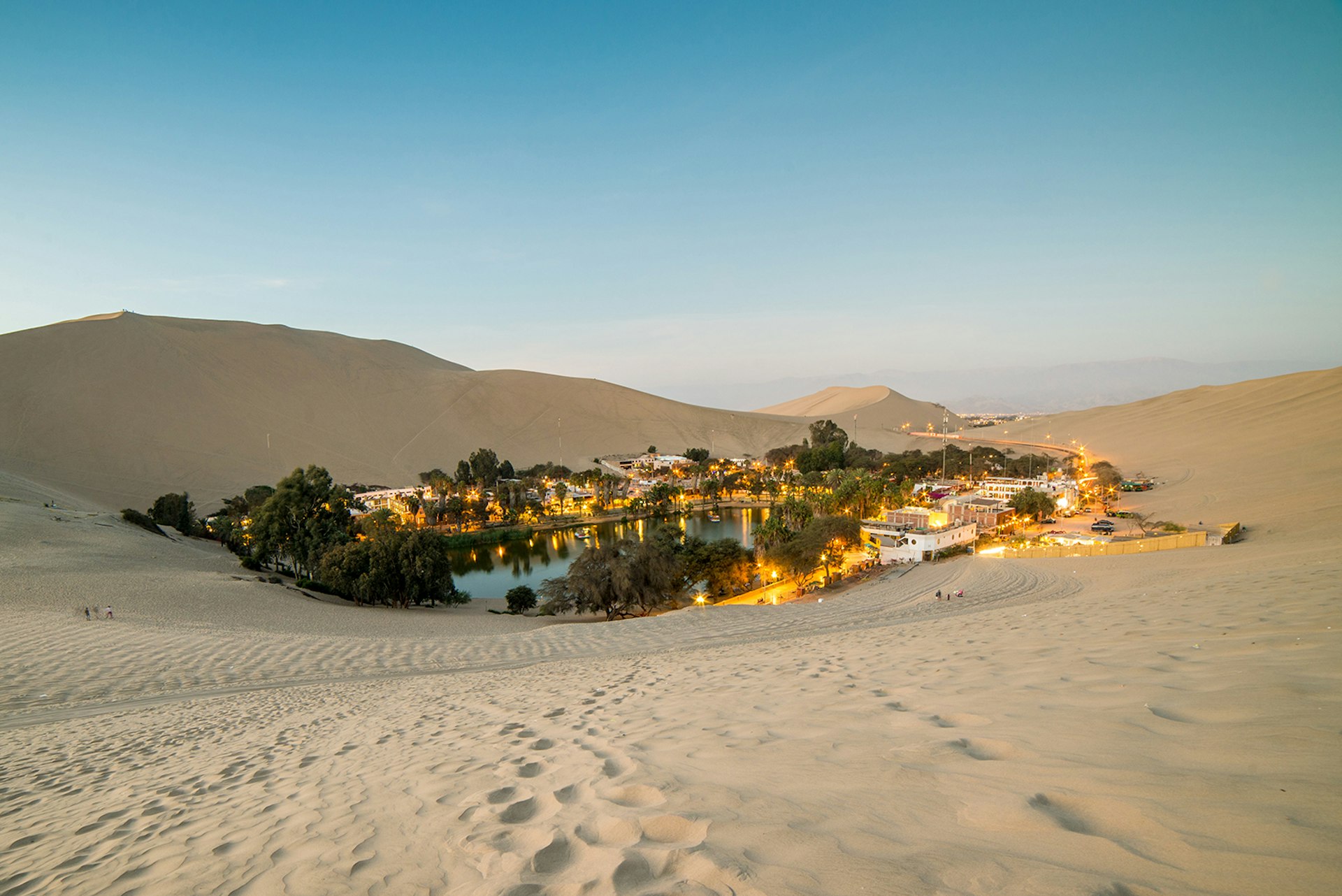 The desert oasis of Huacachina at dusk. The small body of water is surrounded by bushy green trees, white buildings and lights. 