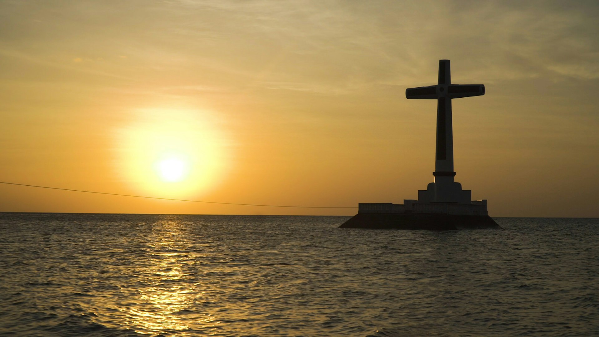A cross juts out from the sea, marking the spot of Camiguin's Sunken Cemetry - one of the Philippines'most unique dive spots. The cross is a silhouette due to the sun setting in the background