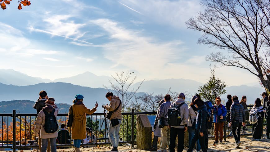 A view from Takao-san: it's a bright autumn day, with red and orange leaves on trees, and shadowy peaks visible in the background; people are in coats in the foreground