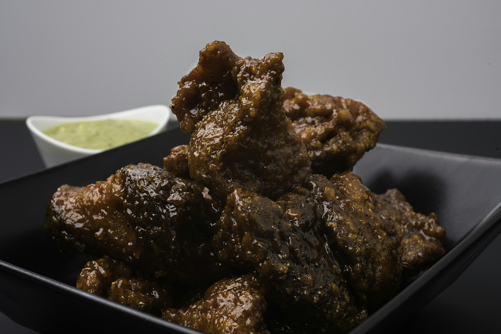 A pile of dark, sauced vegan chicken wings sits on a plate, with a bowl of yellow sauce in the background