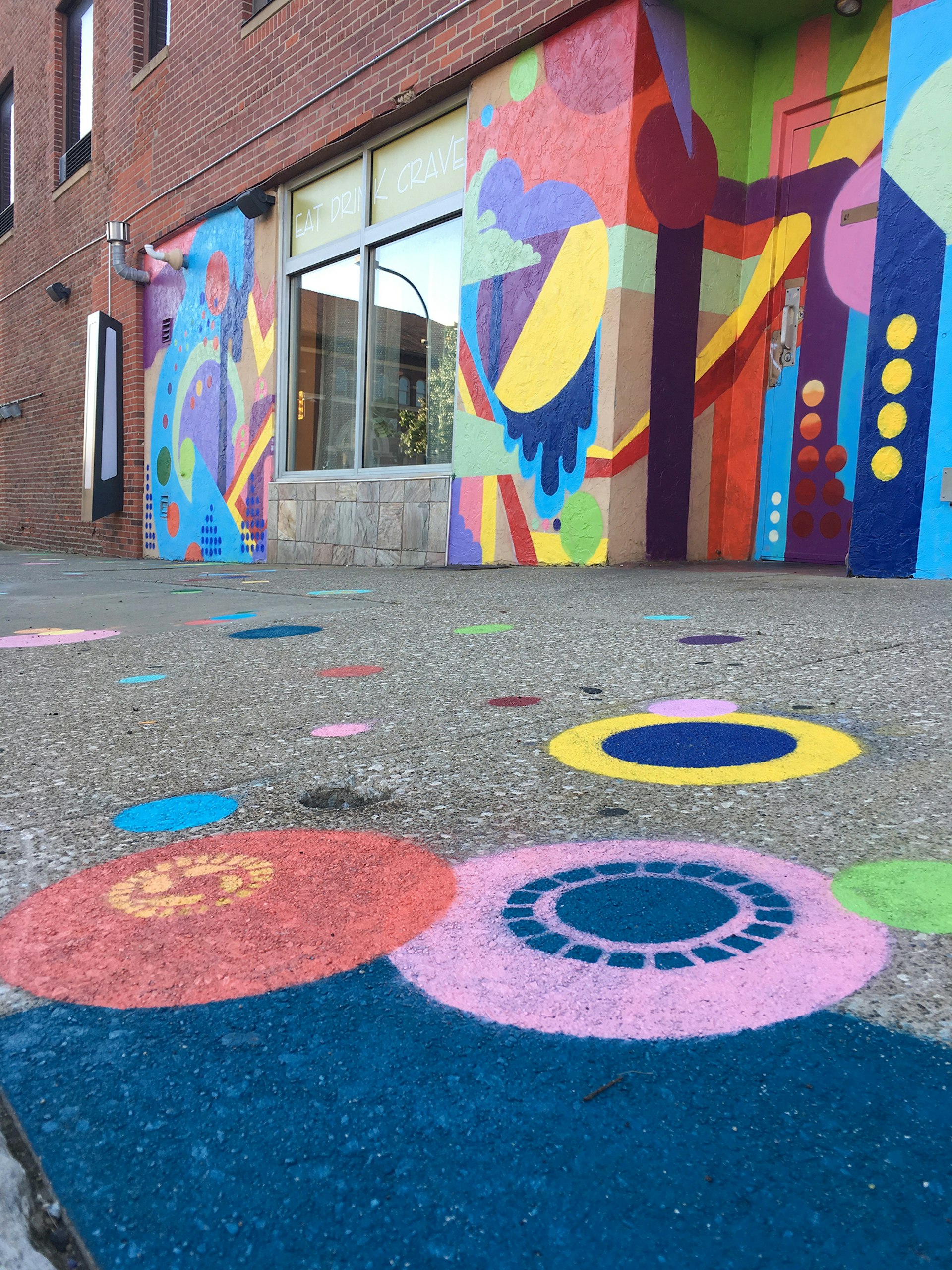 An old brick building is now covered in abstract geometric designs in yellow, blue, purple, orange and red. The sidewalk in front of building has colorful circles painted on it.