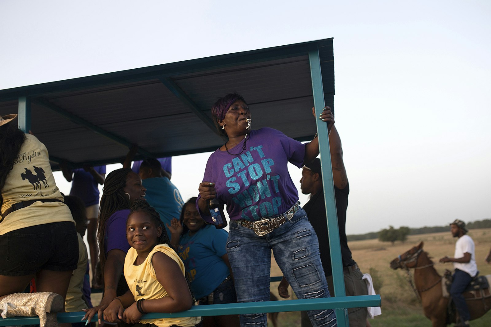 group of women, one in a purple shirt that says 'can't stop won't stop' dance on the back of a covered trailer, with horses in the background, on a trail ride