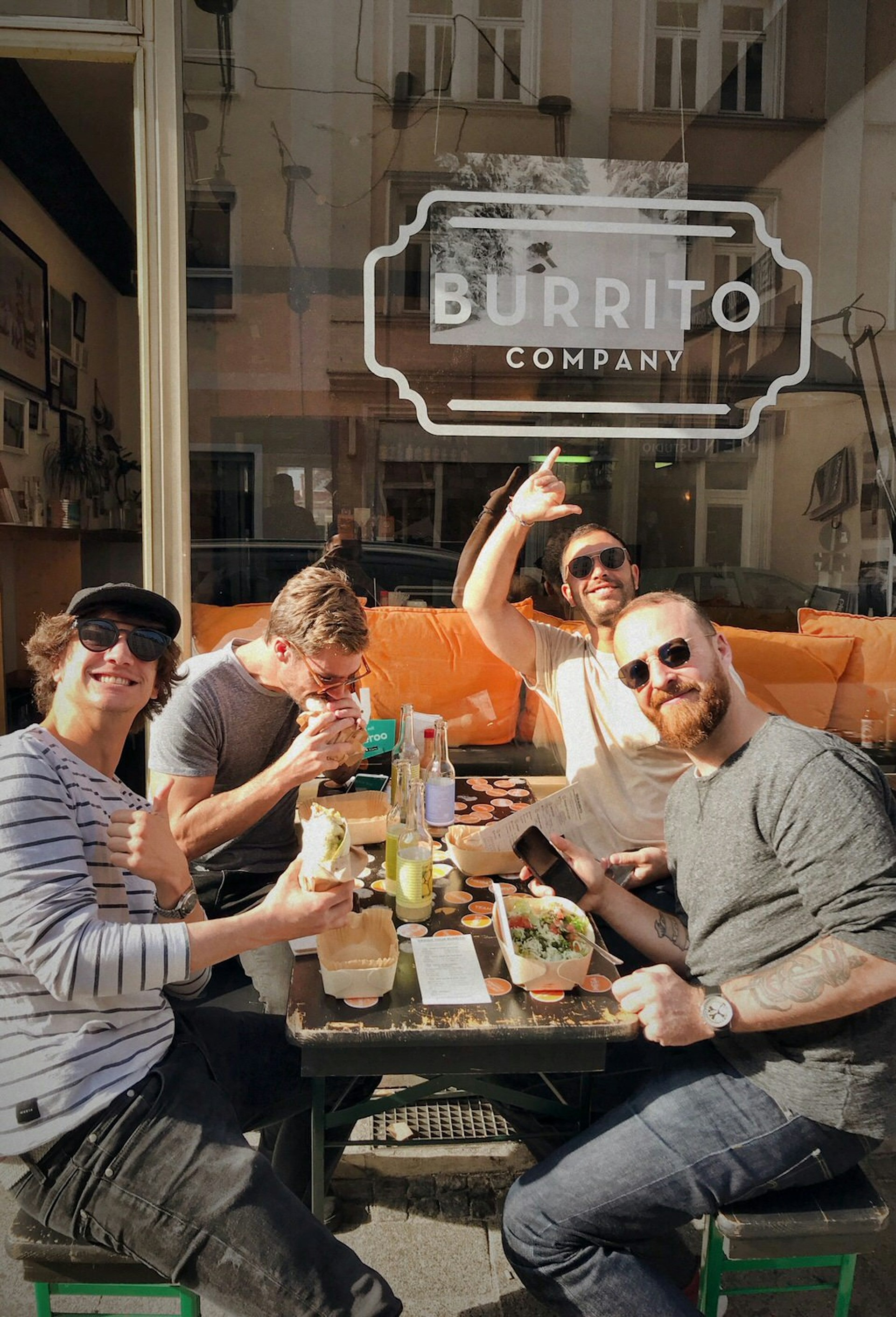 A group of men sitting at a table in the su outside the Burrito Company. The table has a mix of Mexican food. 
