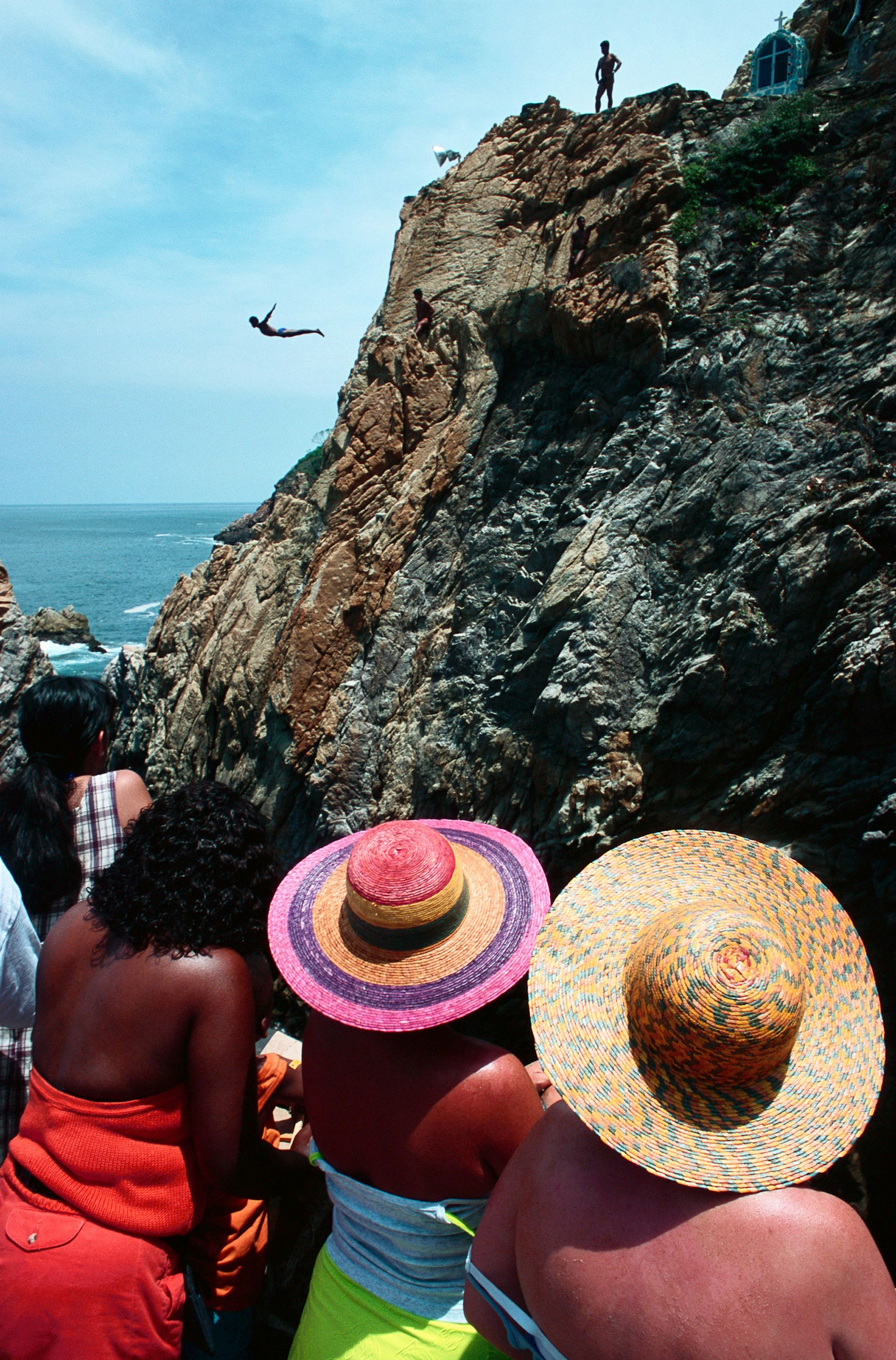 Three women in sun hats watch men dive from a high cliff into the ocean