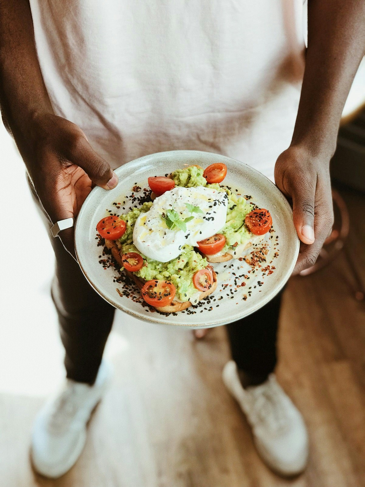 Someone holding a plate of beautifully presented avocado toast topped with burrata cheese, sprinkled with black pepper and surrounded by vibrant red tomatoes © Mary's Coffee Club
