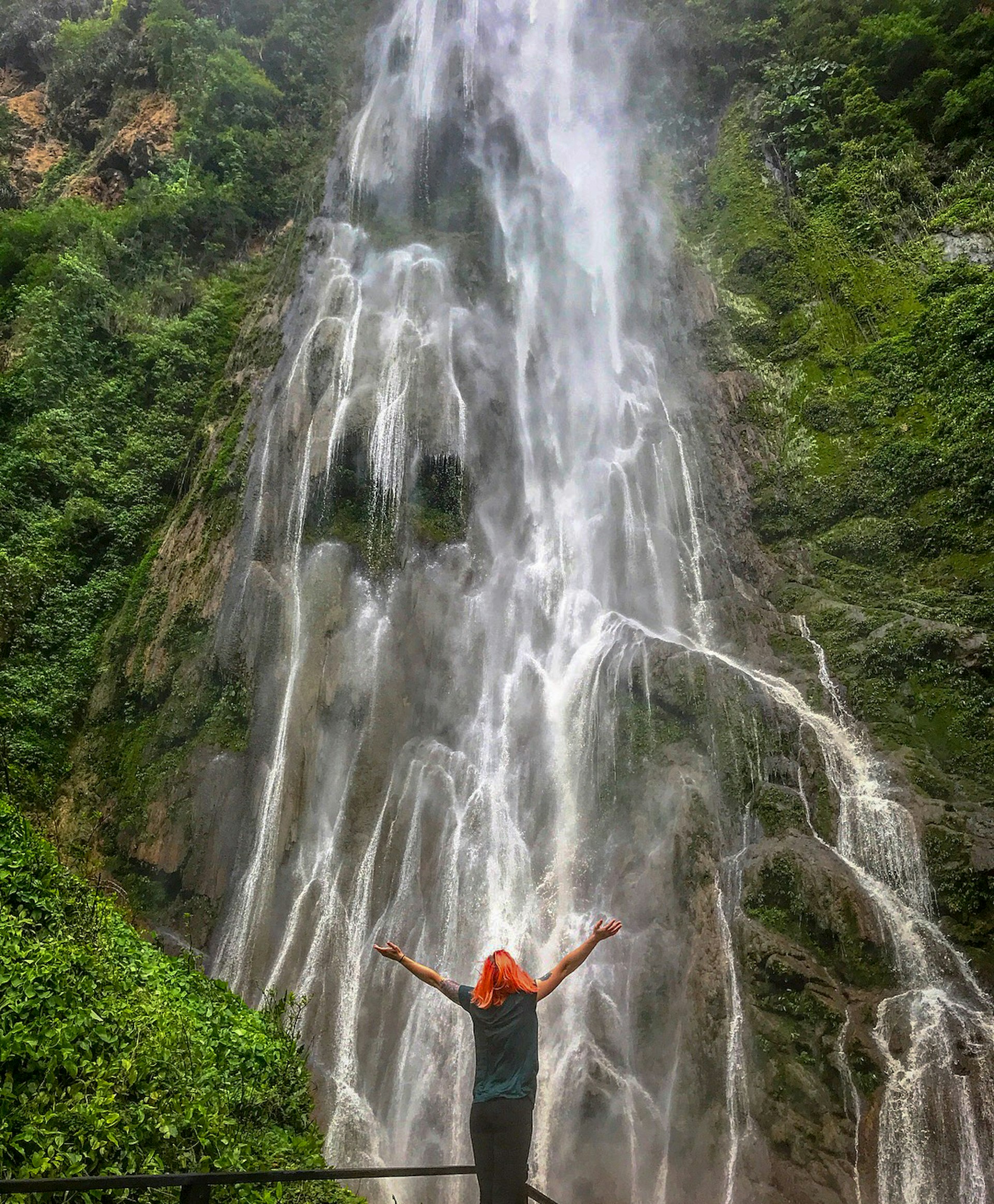 A woman with orange hair stands in front of a tall waterfall with her arms outstretched