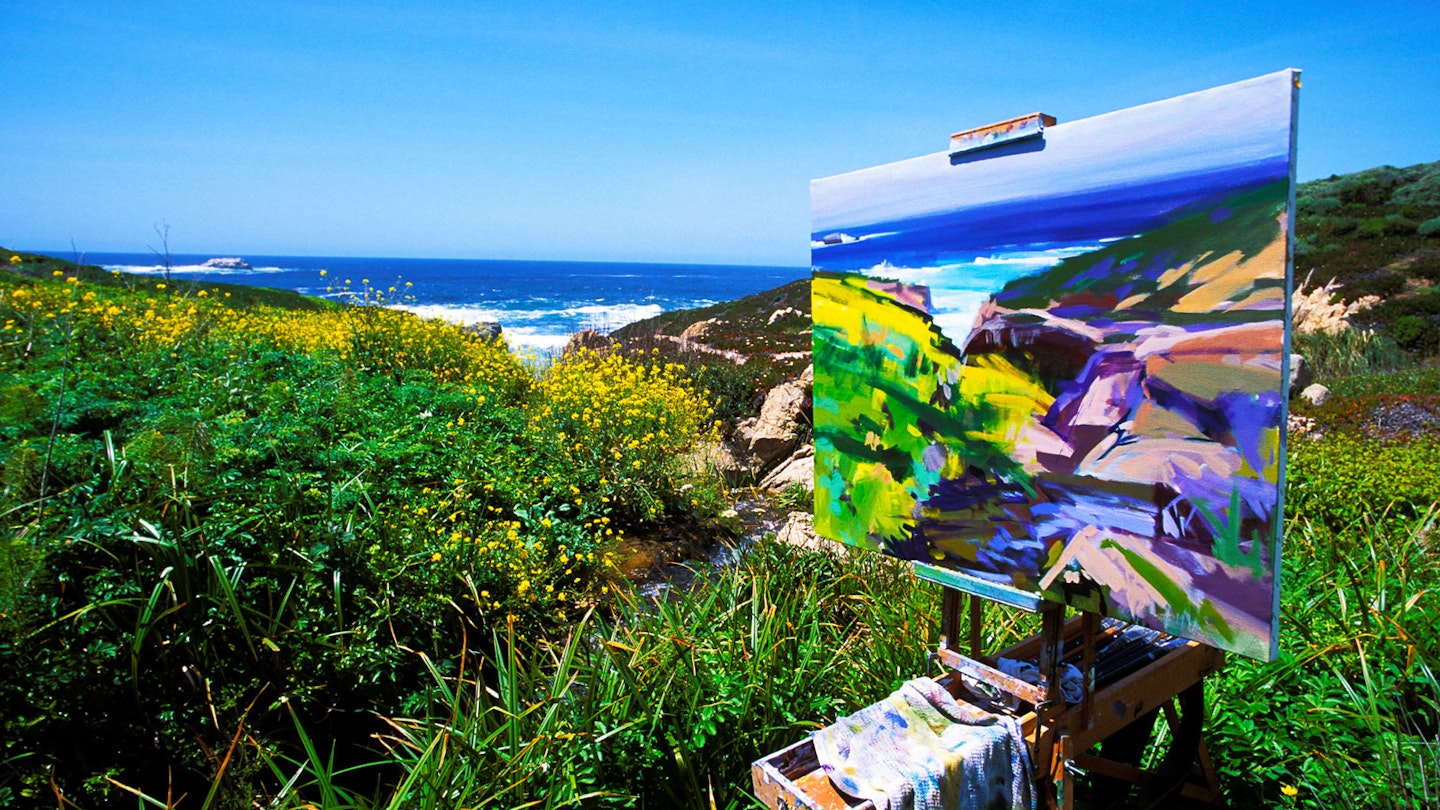 a finished landscape stands on an easel in with wildflowers and ocean in the background
