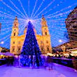 A tall Christmas tree covered in small blue lights stands in the middle of an ice rink in front a large church. Strings of lights fan out from the top of the tree to form a canopy of lights above the ice skaters