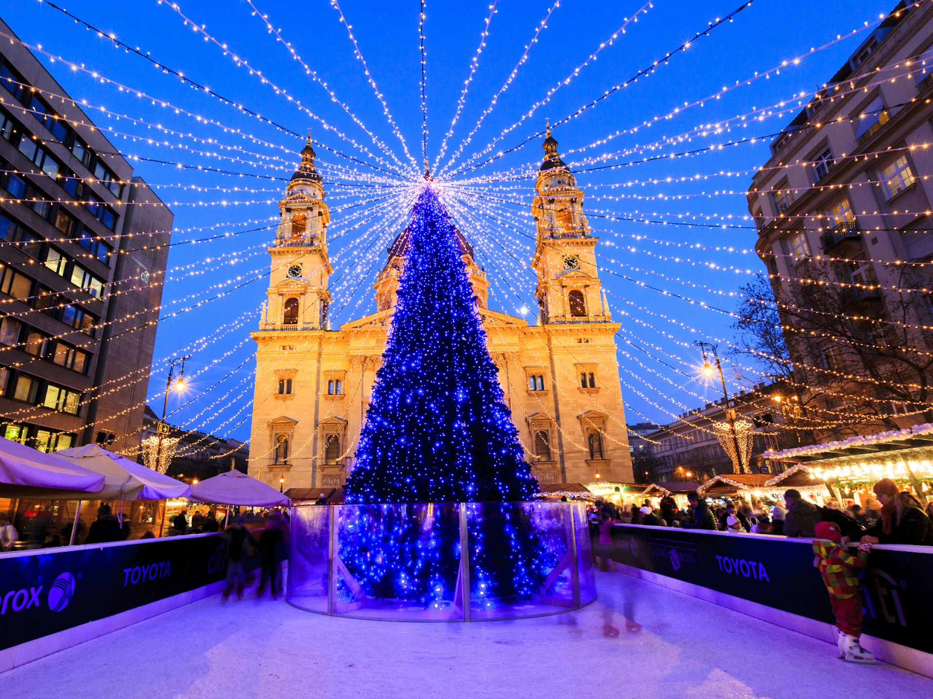 A tall Christmas tree covered in small blue lights stands in the middle of an ice rink in front a large church. Strings of lights fan out from the top of the tree to form a canopy of lights above the ice skaters