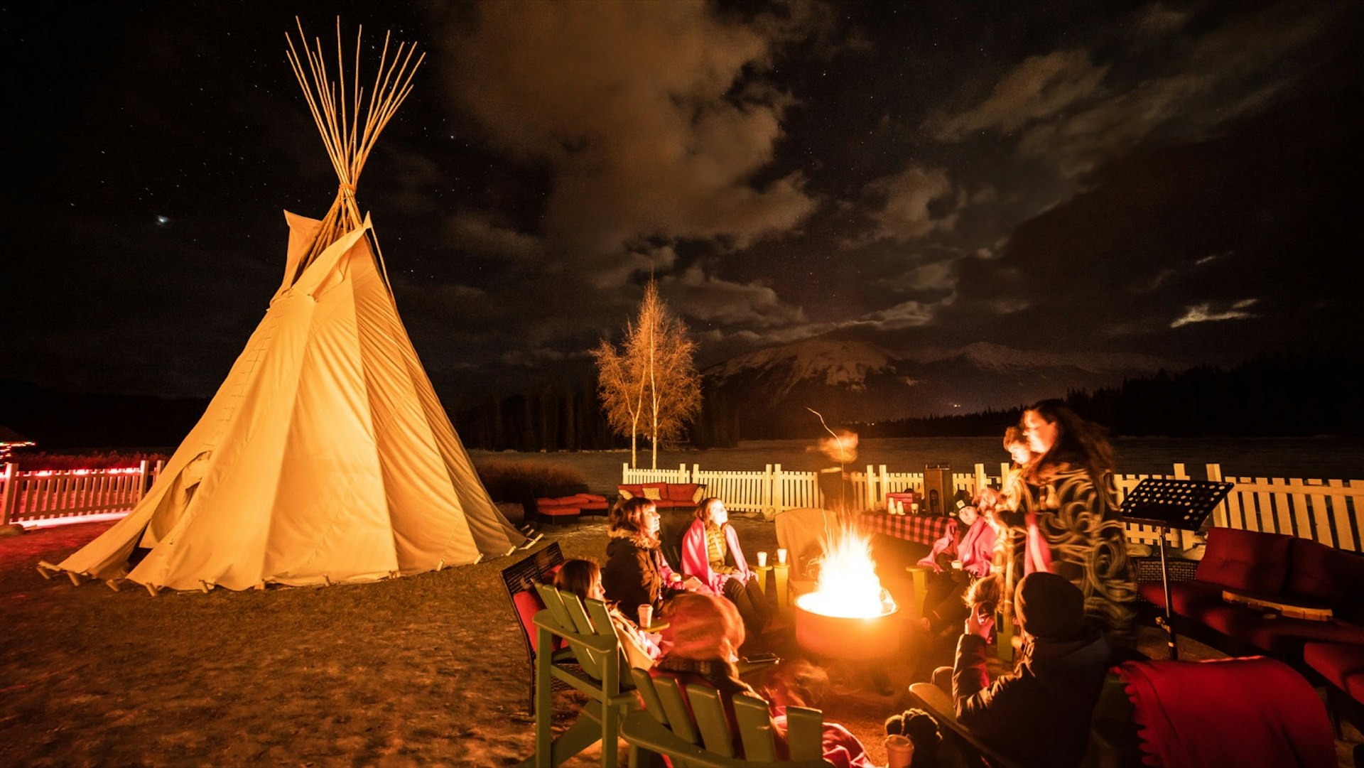 A tipi sits nearby as a group of people talk around a campfire and look up at a sky full of stars; Alberta Canada winter adventures