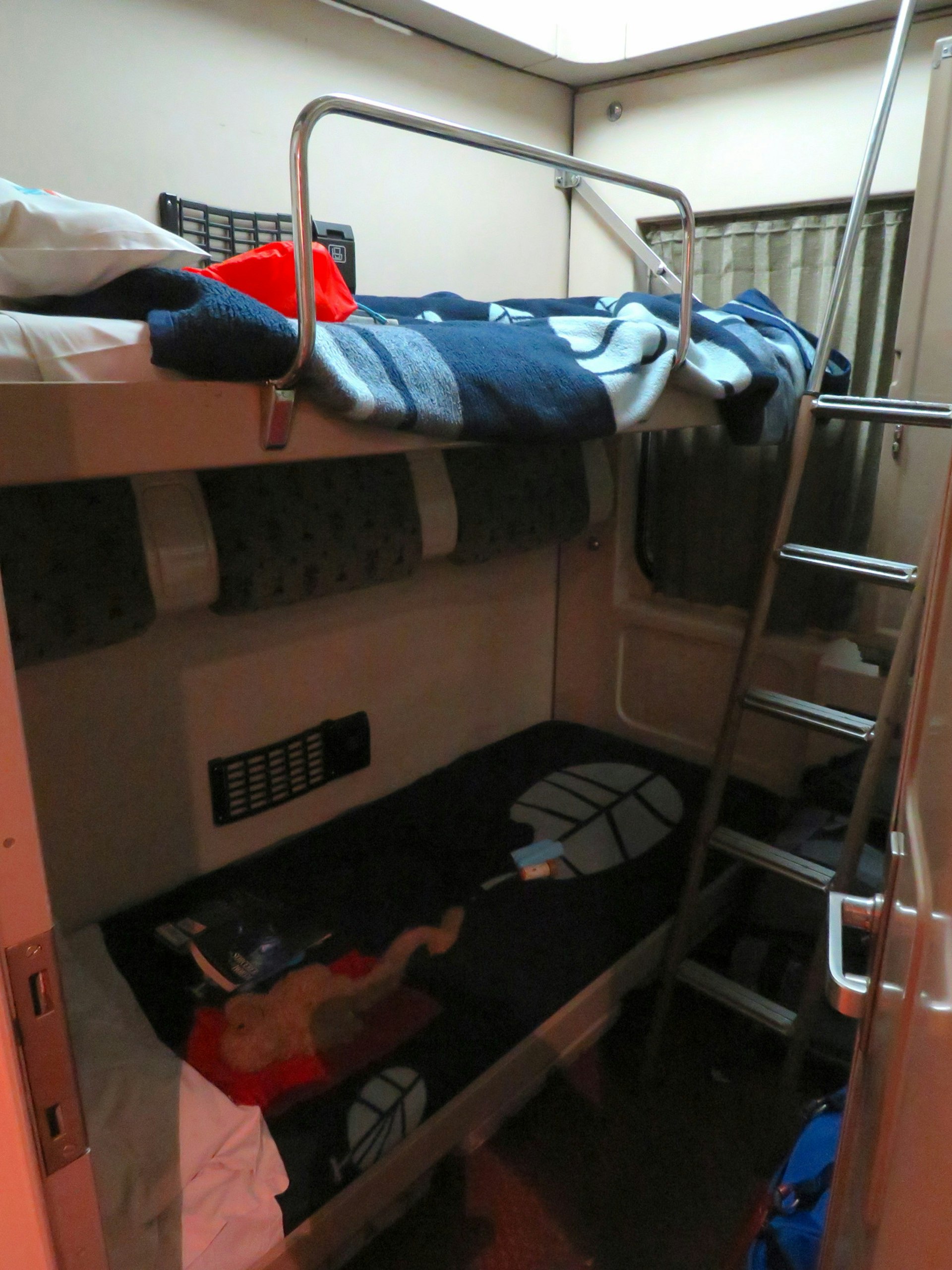 Two bunks in a small compartment on a train with a stainless steel ladder at the end