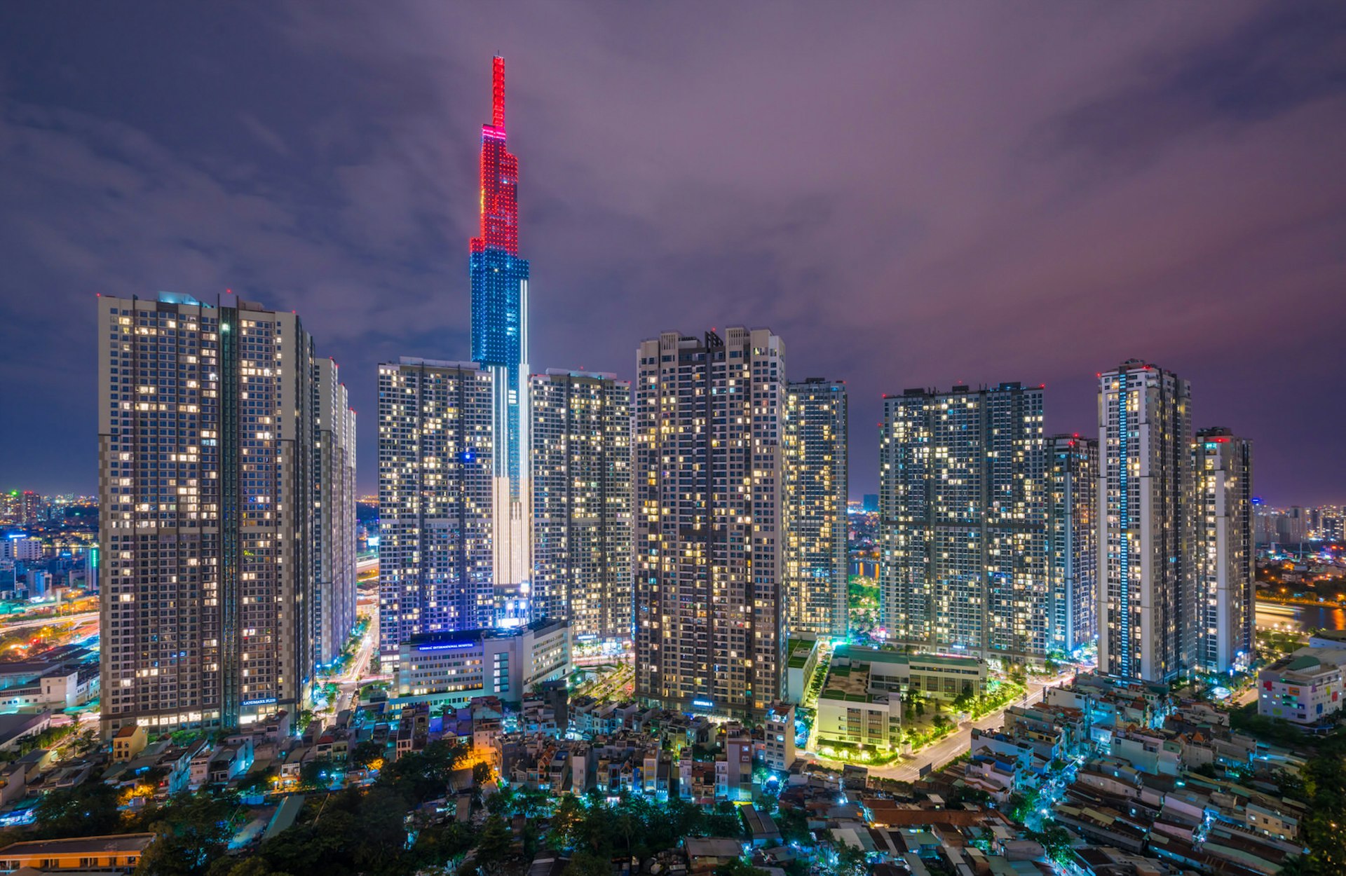 Landmark 81, pictured at night, towers above Ho Chi Minh City. The skyscraper is illuminated by white, blue and red lights in horizontal bands and surrounded by smaller, sparkling buildings. 