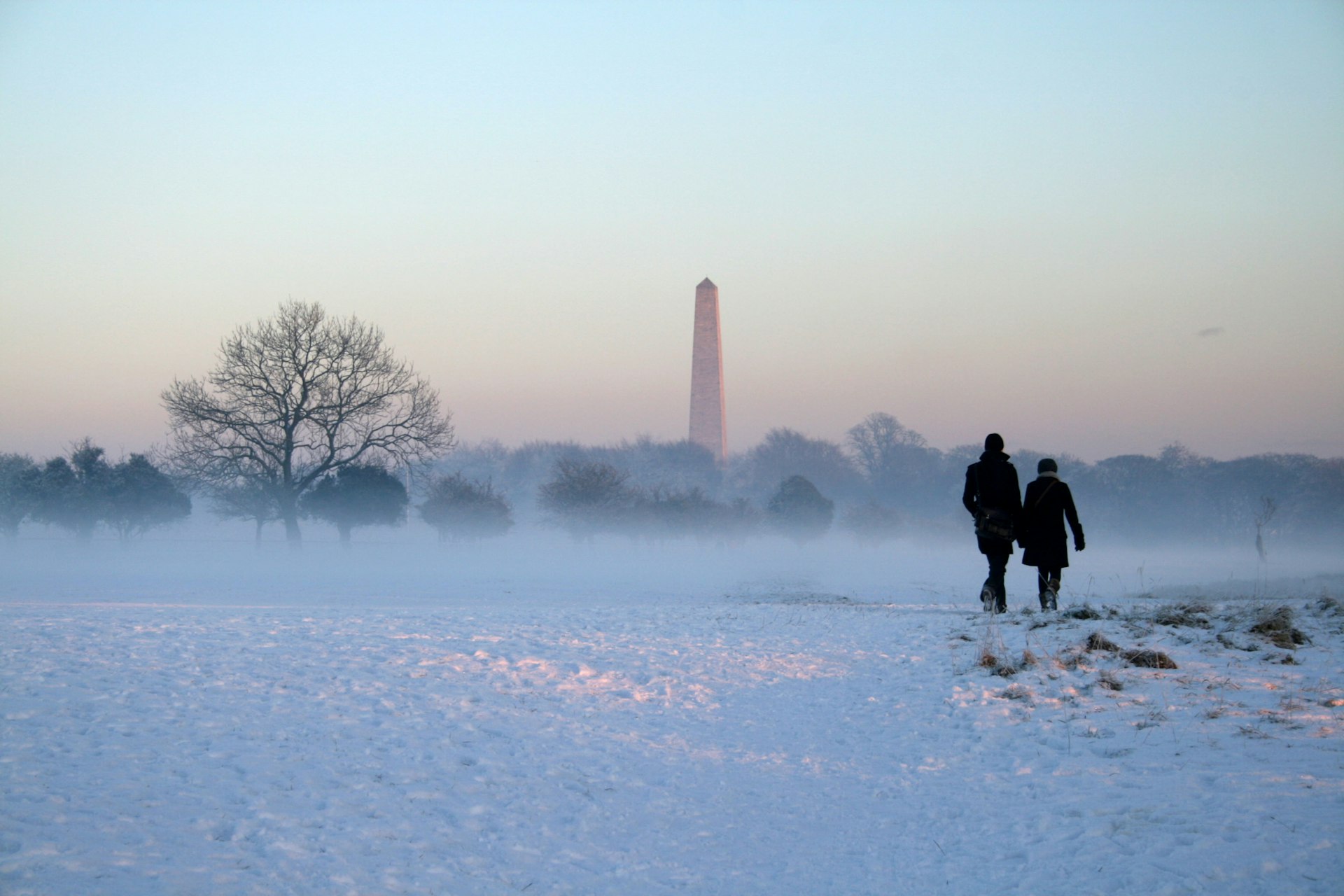 Phoienix Park Dublin in winter. Two people walk across the park which is covered in snow towards a pink horizon and a lone stone tower. 