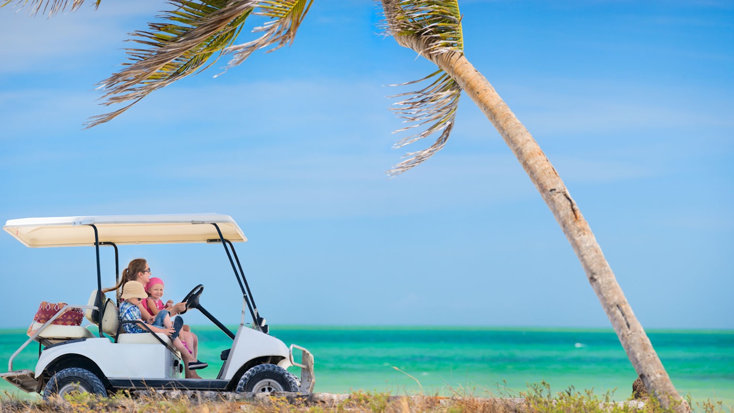 a woman drives a golf cart with a young girl on her lap and a boy next to her on the beach toward a palm tree